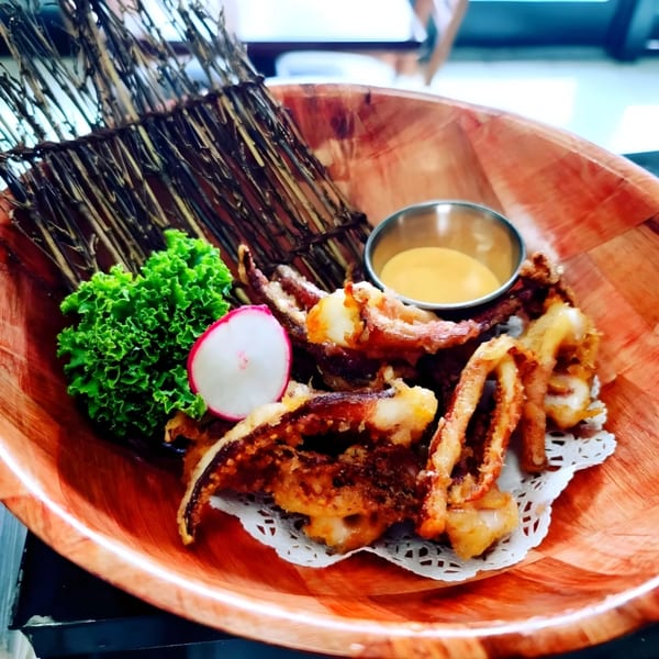 Crispy fried calamari with a side of sauce, lettuce, and radish in a wooden bowl.